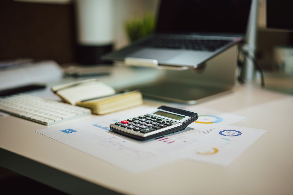 Black calculator resting on printed financial charts next to a white keyboard and notebook